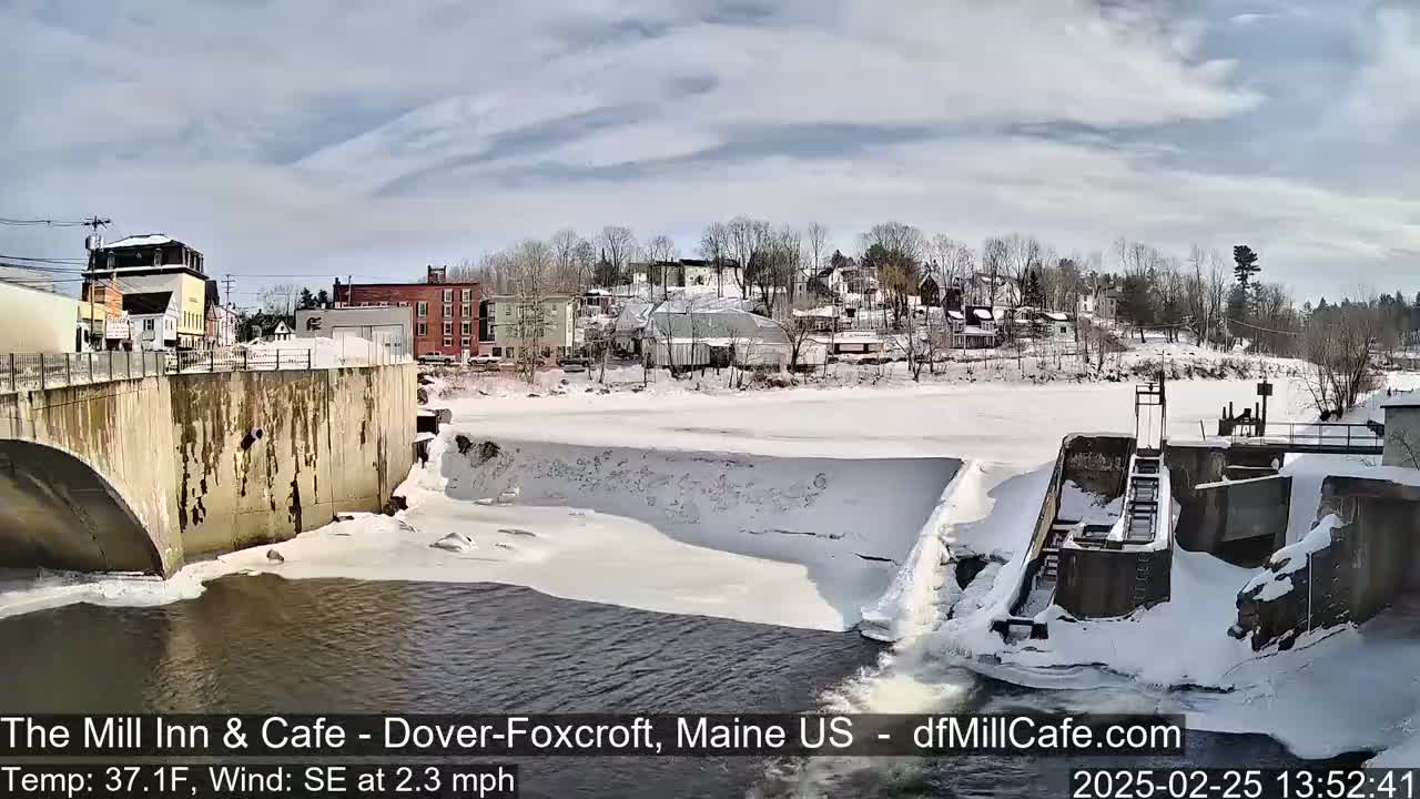 A partially frozen river flows past a snow-covered dam and town under a partly cloudy sky.