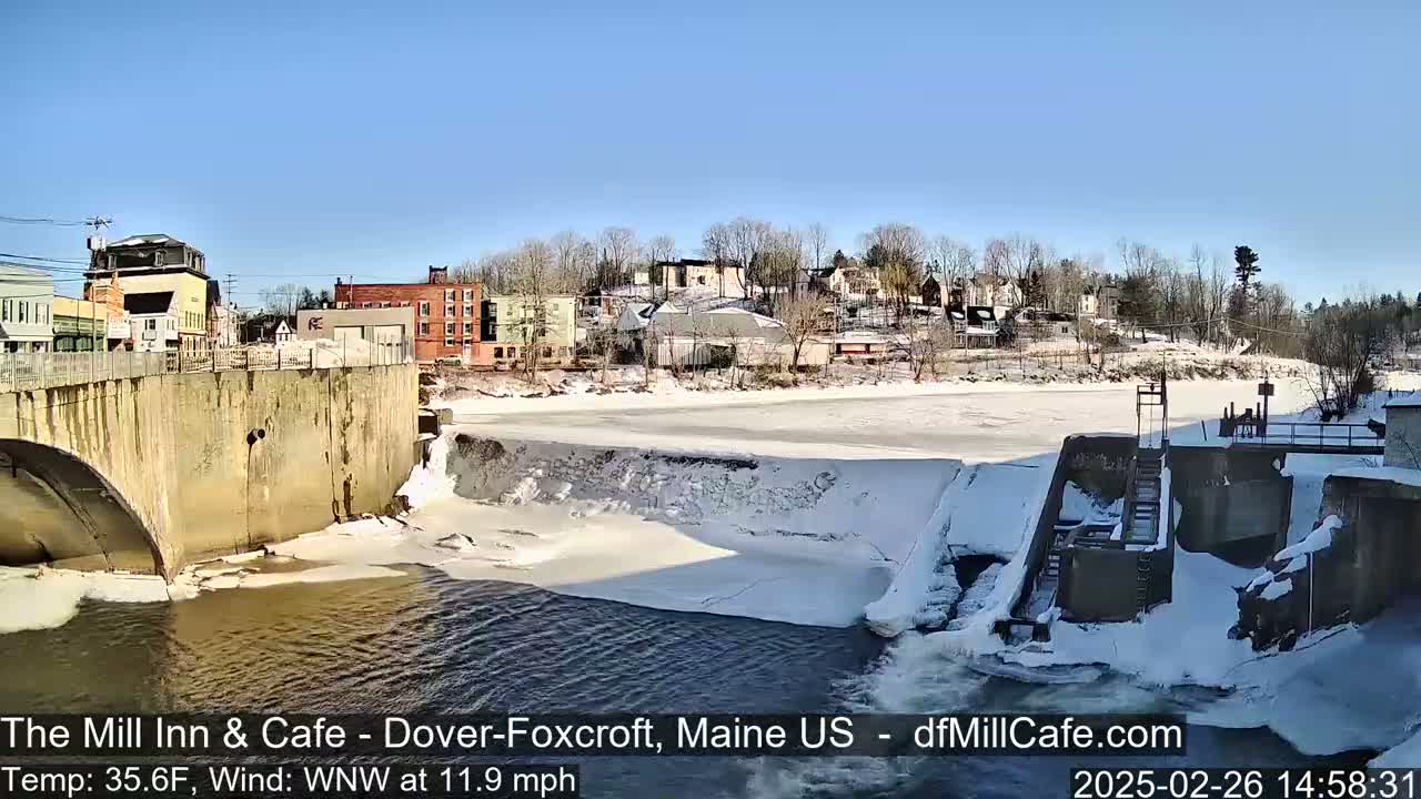 A partially frozen river flows past a dam and a town under a clear, sunny sky.