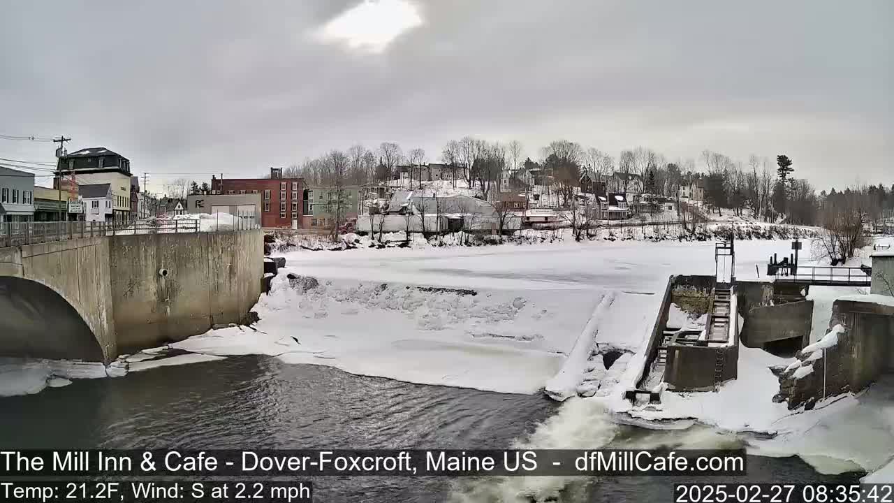 A partially frozen river flows past a snow-covered dam and town under an overcast sky.