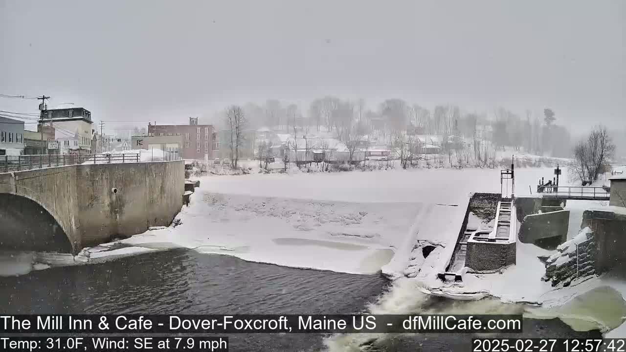 A snow-covered river flows past a dam and under a bridge in a small town during a snowfall.