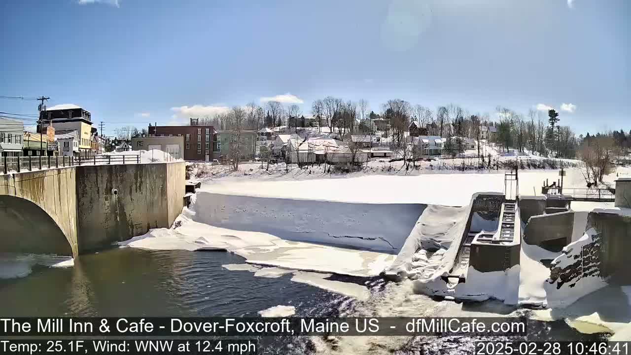 A snow-covered dam and river flow past a small town under a bright, sunny sky.