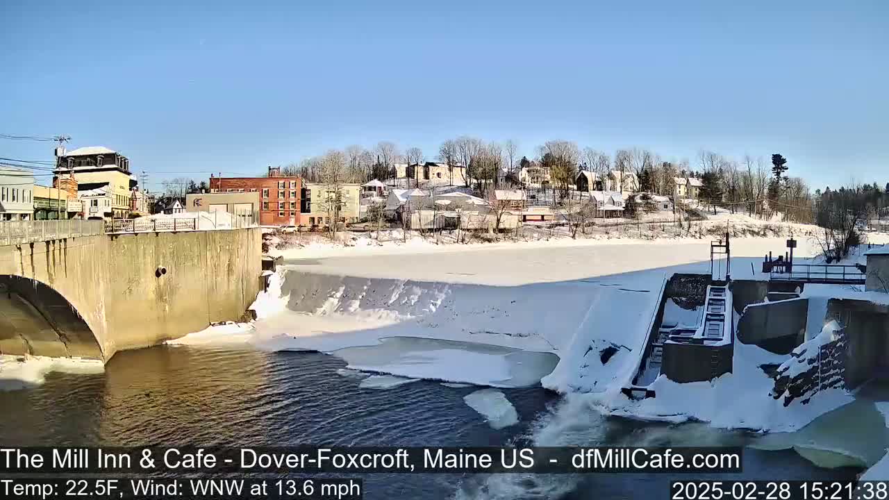 A partially frozen river flows past a snow-covered dam and town under a clear blue sky.