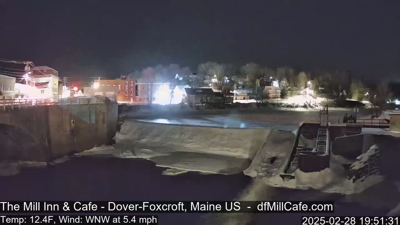 A nighttime view of a snow-covered river flowing past a dam and buildings under a dark sky.