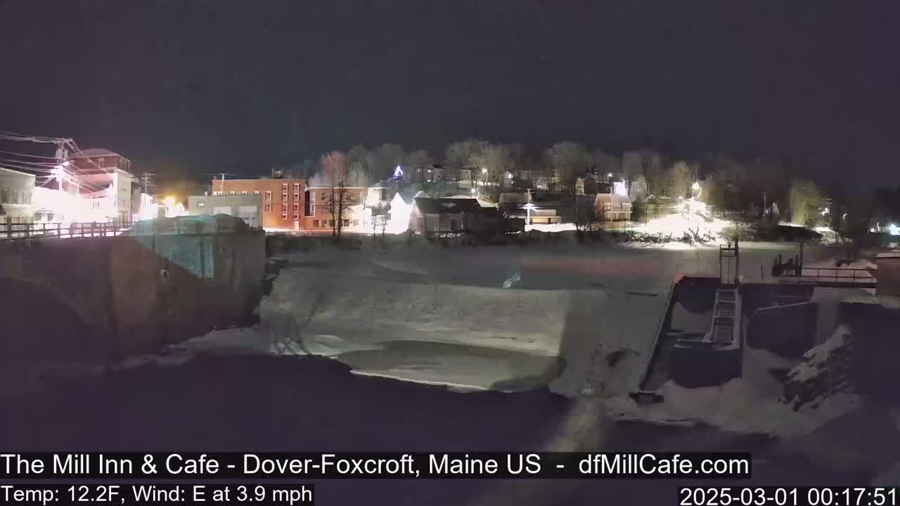 A snow-covered town at night, viewed from a bridge overlooking a partially frozen river and a dam.