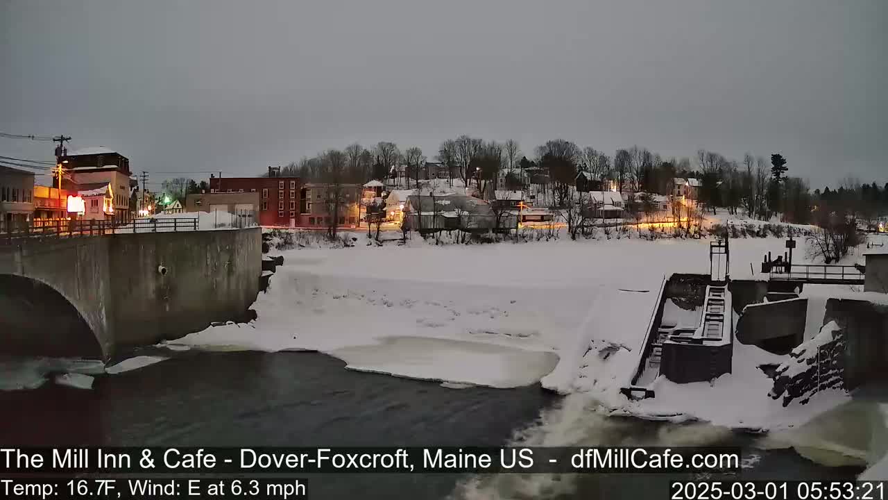 A snow-covered town is seen at dusk, with a river partially frozen and flowing past a dam and under a bridge.