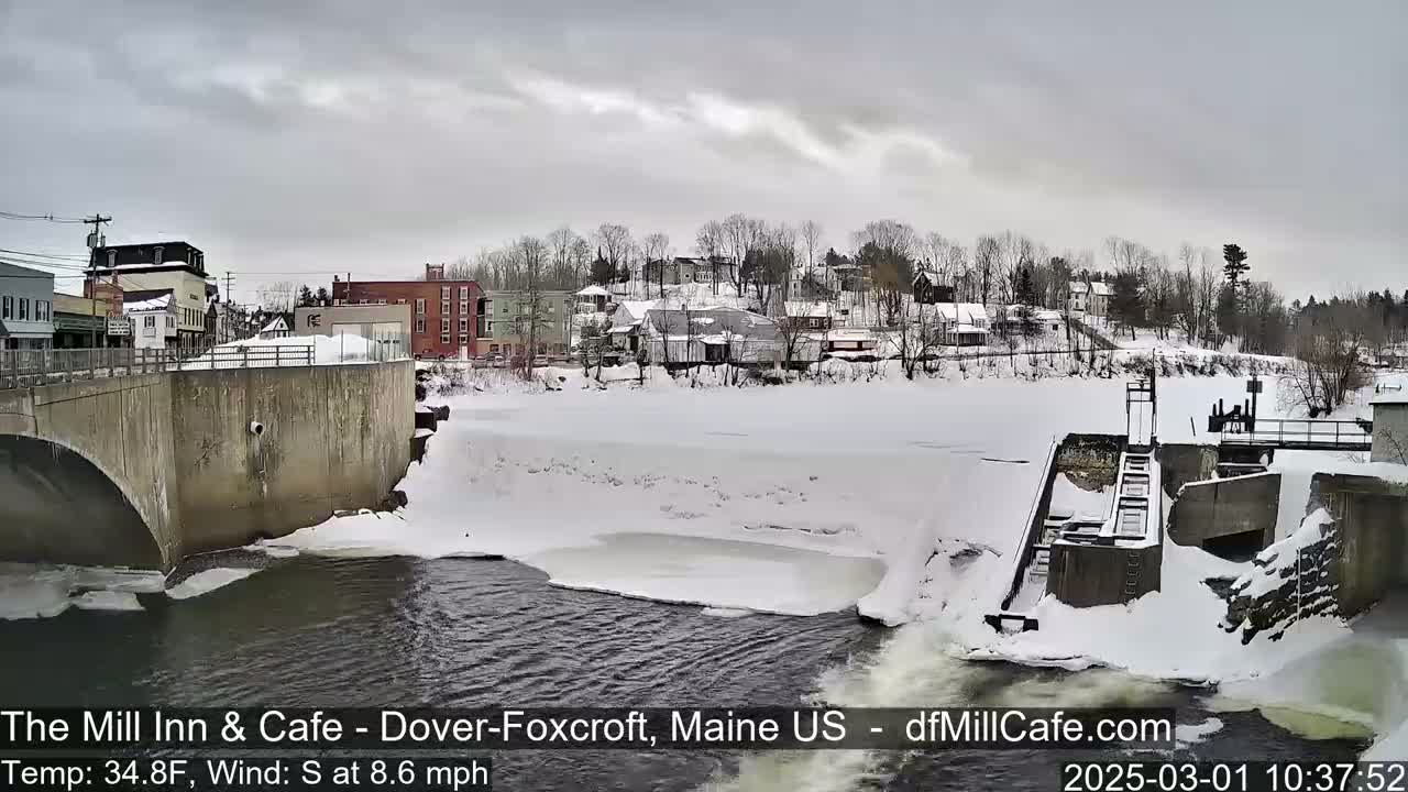 A partially frozen river flows beneath a bridge past a snow-covered dam and a small town under a cloudy sky.