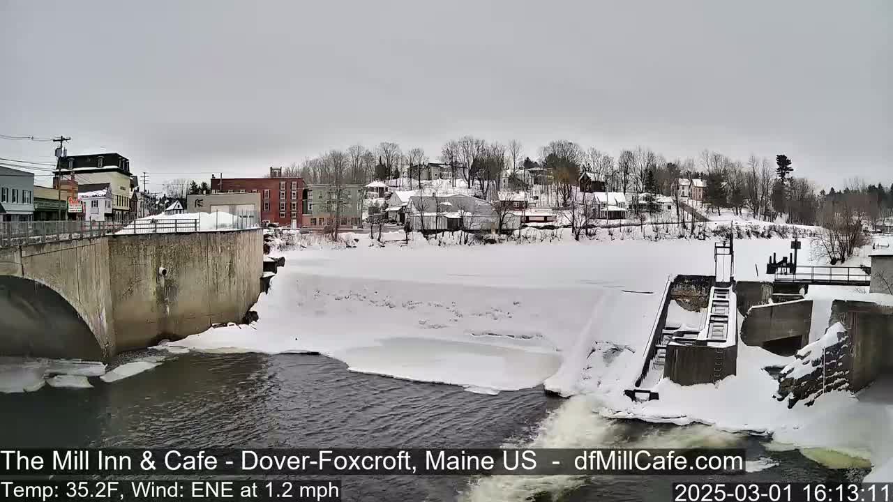 A snow-covered town is seen from a bridge overlooking a partially frozen river with a dam and a low-flow waterfall, under an overcast sky.