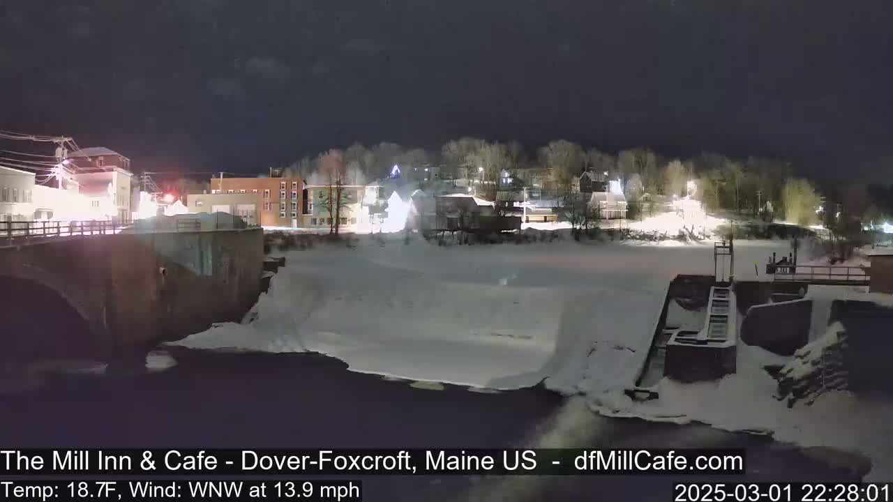 A snow-covered town at night is seen from across a partially frozen river, with a dam visible in the foreground.