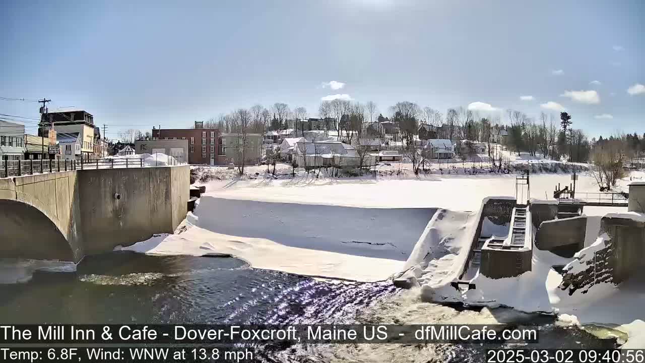 A snow-covered town is viewed from a bridge overlooking a partially frozen river with a dam and water flowing over it under a sunny sky.