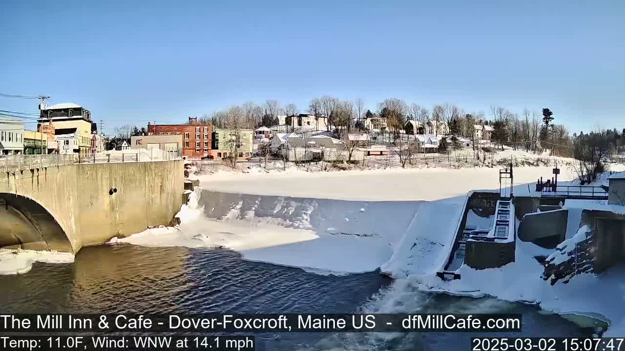 A partially frozen river flows under a bridge next to a snow-covered town on a sunny, cold day.