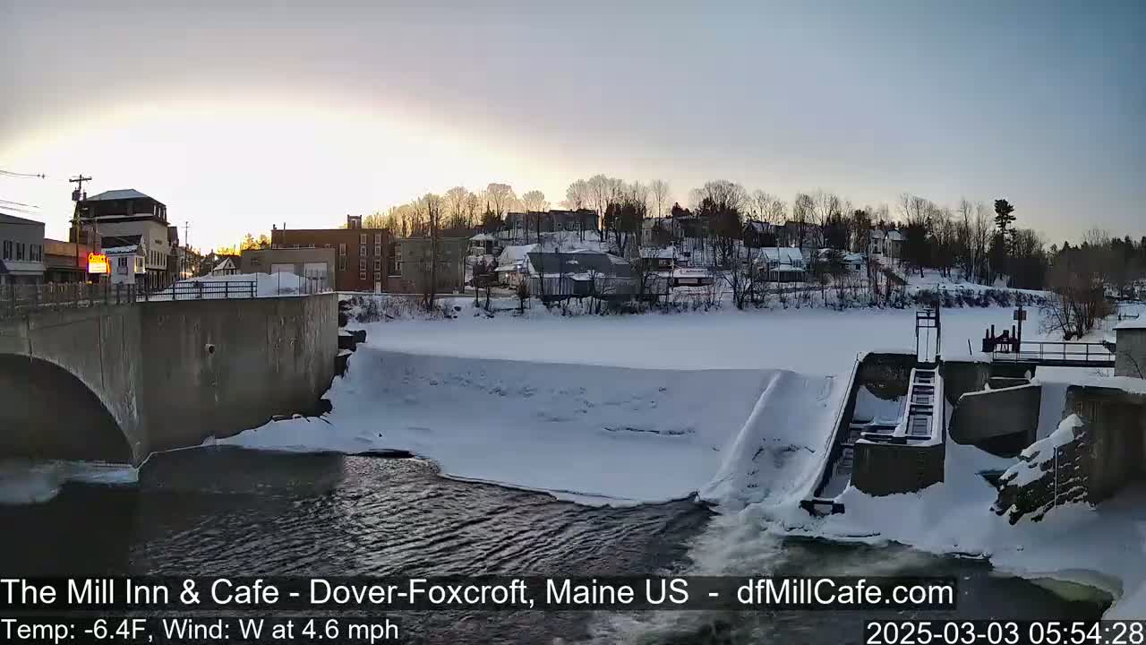 A snow-covered town is seen from a bridge overlooking a partially frozen river with a dam and lock system, under a partly cloudy sky.