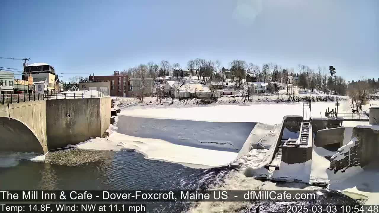 A partially frozen river flows past a dam and a snow-covered town under a clear blue sky.