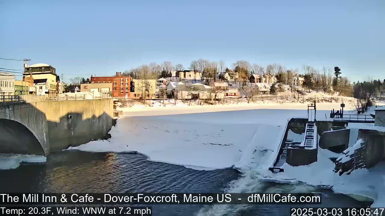 A snow-covered river partially frozen, with a dam and a small town visible in the background under a clear, sunny sky.