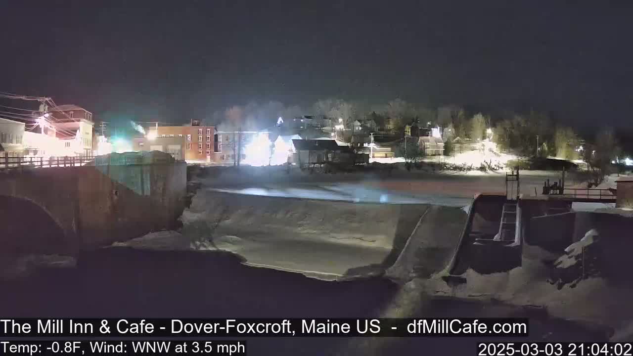 A snow-covered town at night is viewed from a bridge overlooking a partially frozen river, under a clear, dark sky with a wind from the west-northwest at 3.5 mph and a temperature of -0.8°F.