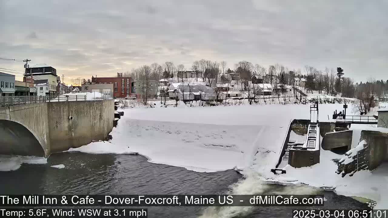 A snow-covered town and river scene with a dam and bridge under a cloudy sky.