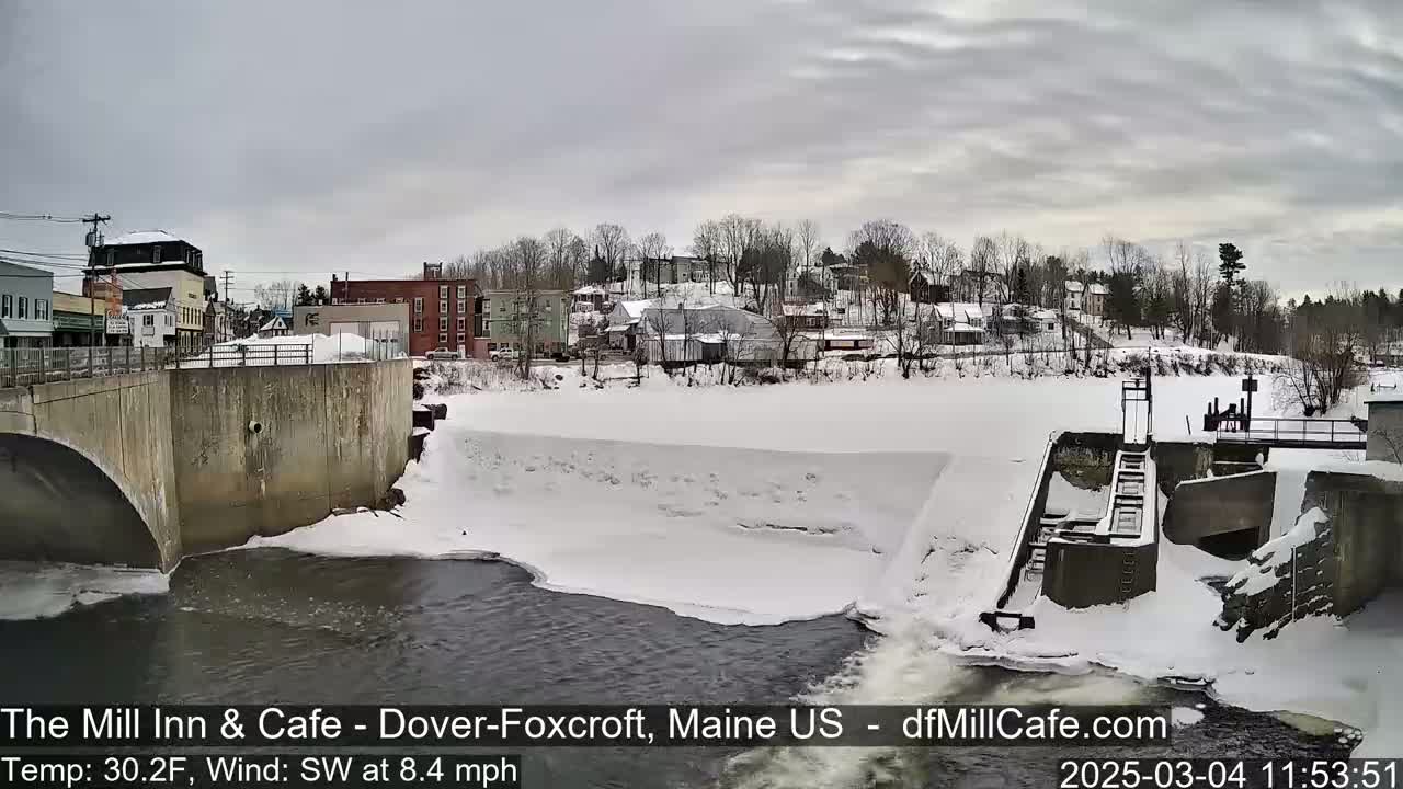 A snow-covered river flows past a dam and a small town under an overcast sky.