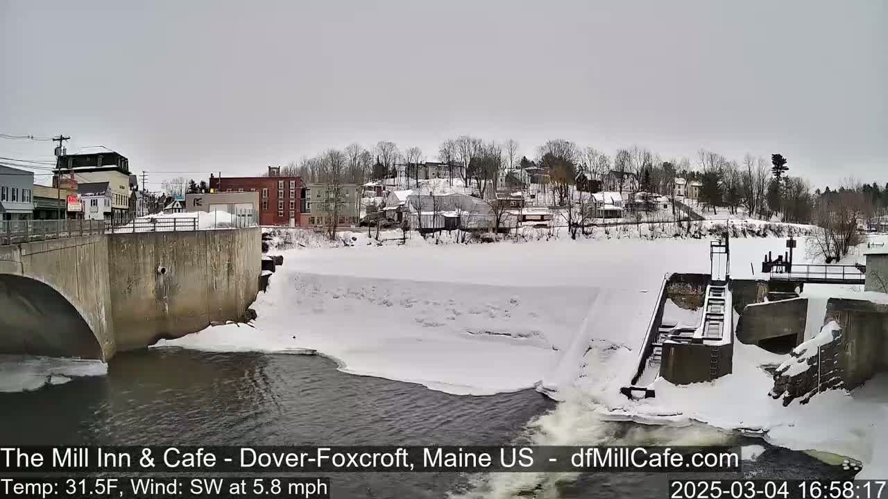 A snow-covered river flows past a dam and under a bridge near a small town on a cloudy day.