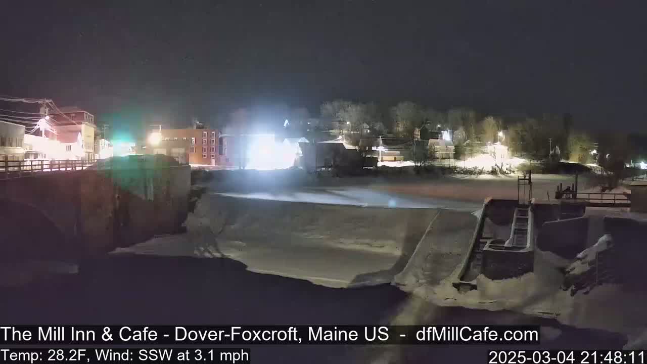 A nighttime view of a snow-covered town by a river with a dam and low light levels, under a clear sky with a temperature of 28.2°F and a south-southwest wind of 3.1 mph.