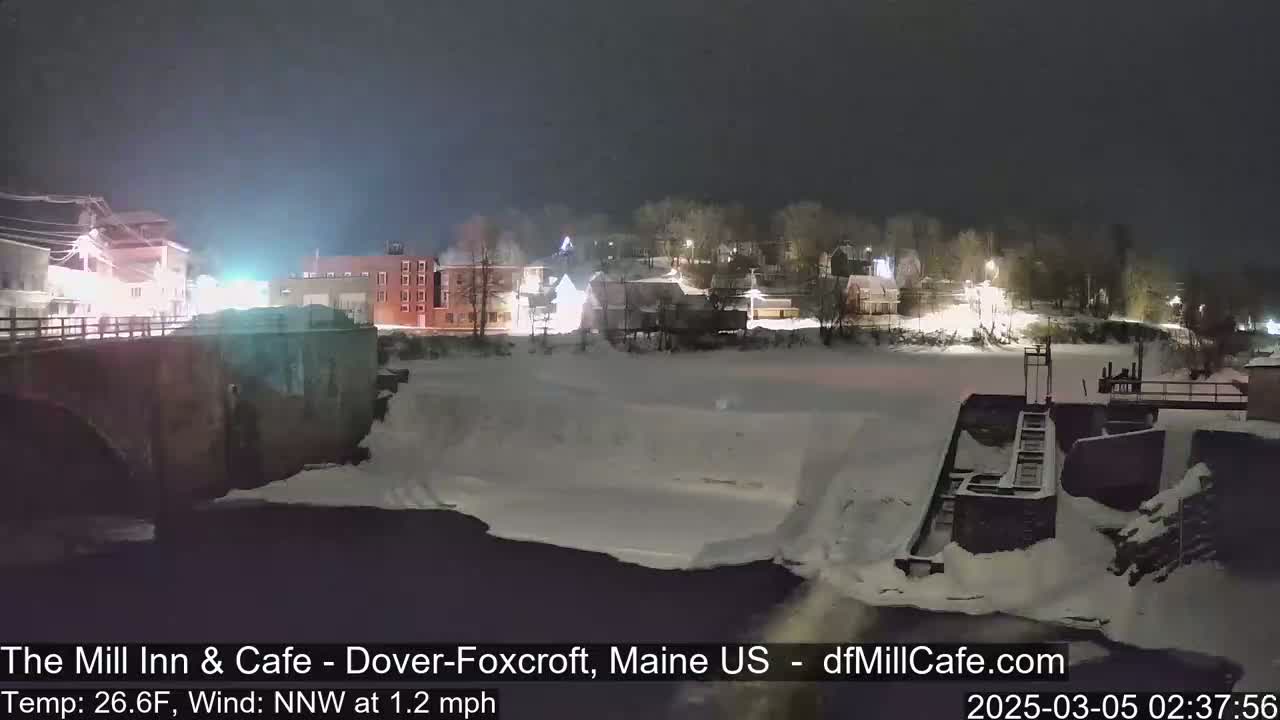 A snow-covered town at night is viewed from a bridge over a partially frozen river, with a light wind.