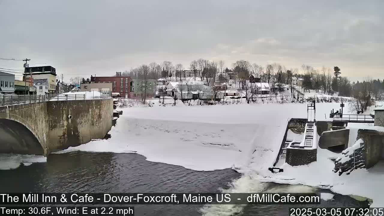 A snow-covered river flows past a dam and a small town under an overcast sky.