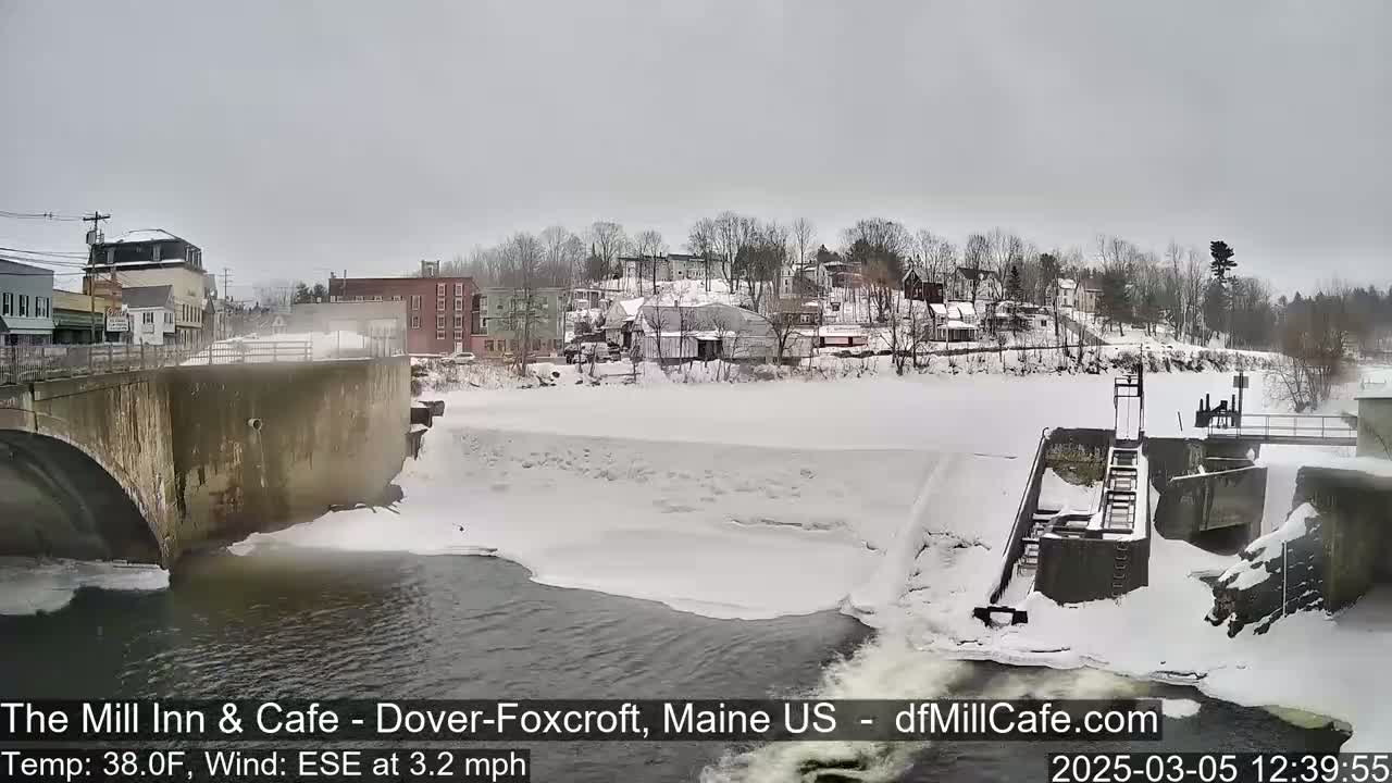 A snow-covered river flows past a dam and under a bridge, with a small town visible in the background under a gray sky.