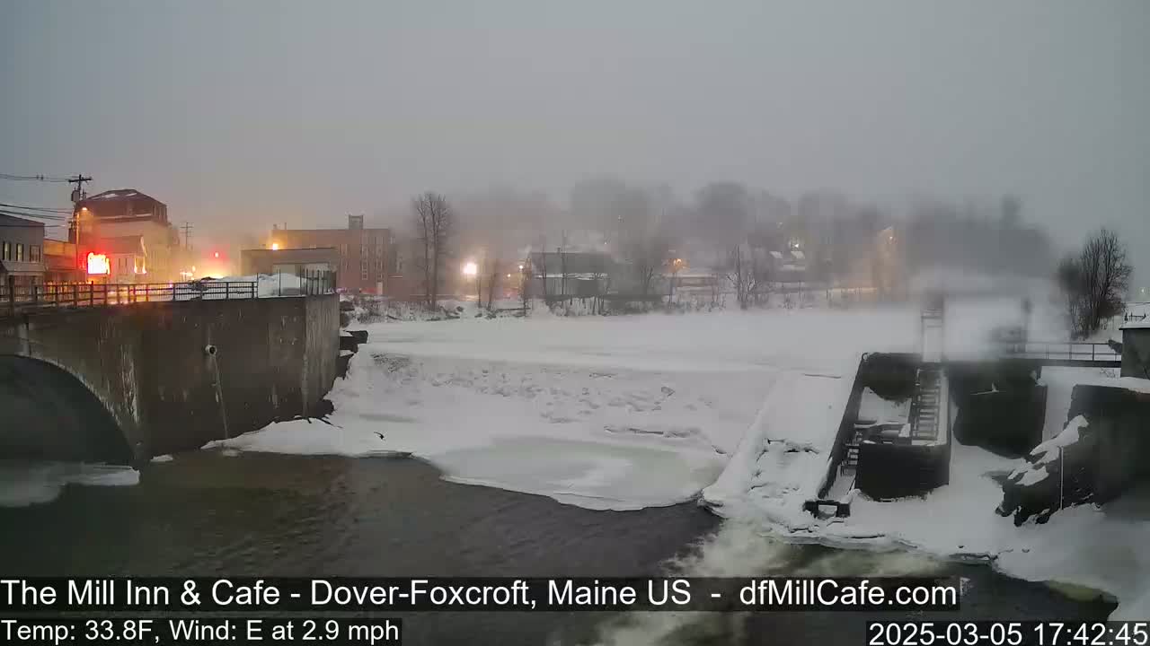 A snow-covered river flows past a bridge and buildings in foggy conditions.