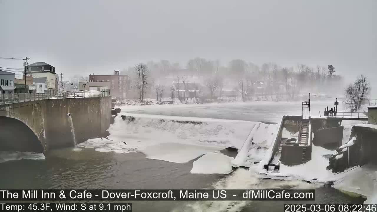 A snow-covered dam and river flow past a town under a foggy, overcast sky.