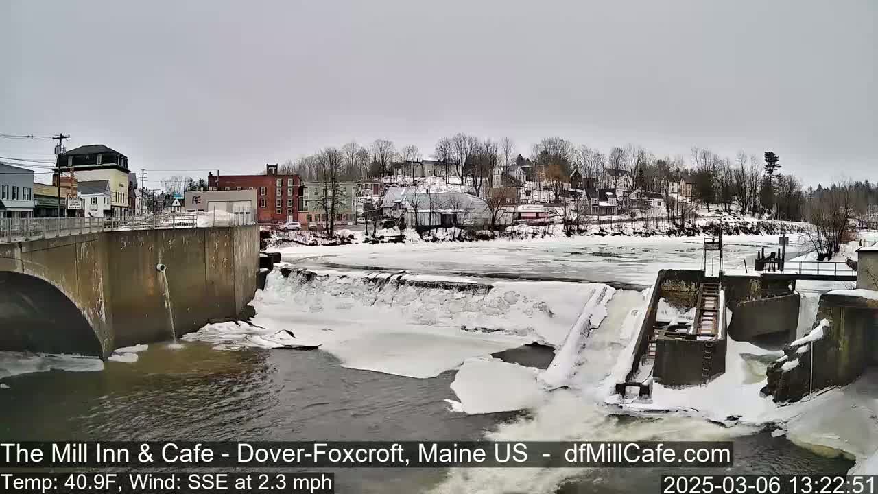 A partially frozen river flows past a dam and a snow-covered town under an overcast sky.