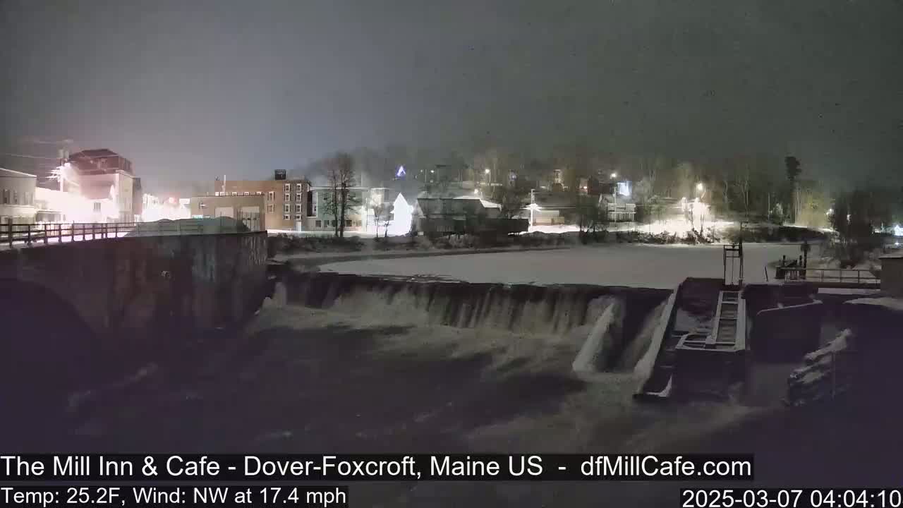 A nighttime view of a snow-covered town with a river flowing over a dam under a cloudy sky.