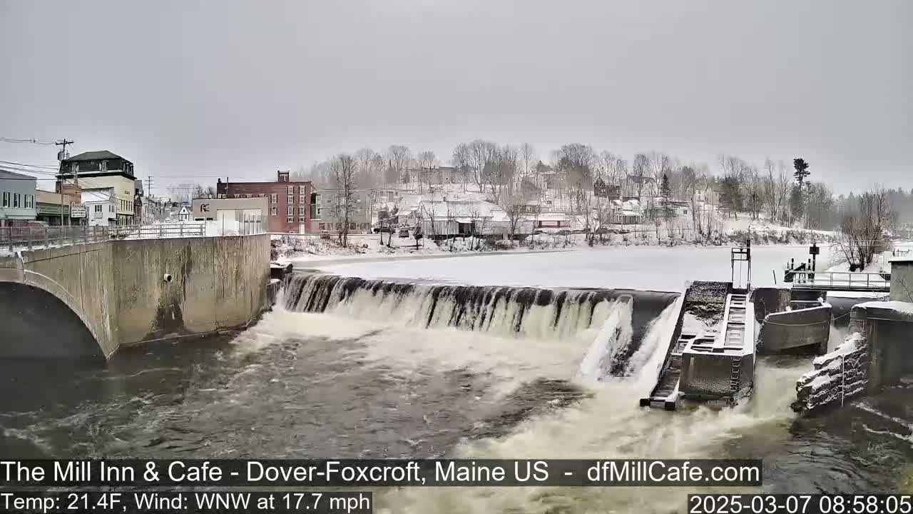 A snow-covered town sits beside a river flowing over a dam under a cloudy sky.