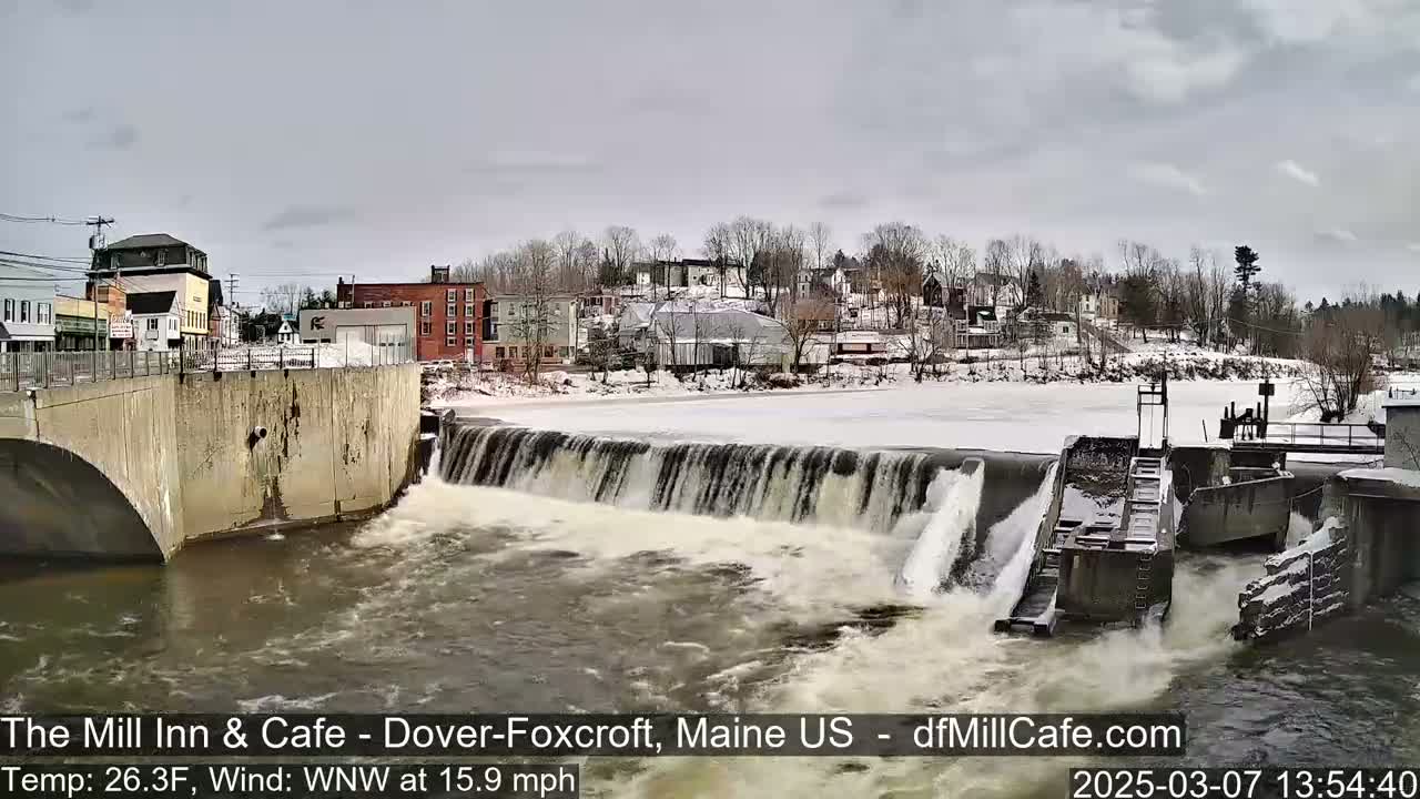 A partially frozen river flows over a dam in a small town under an overcast sky.