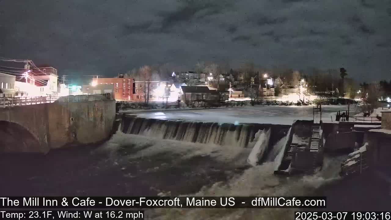 A nighttime view of a dam and waterfall flowing past a town under a dark, cloudy sky.