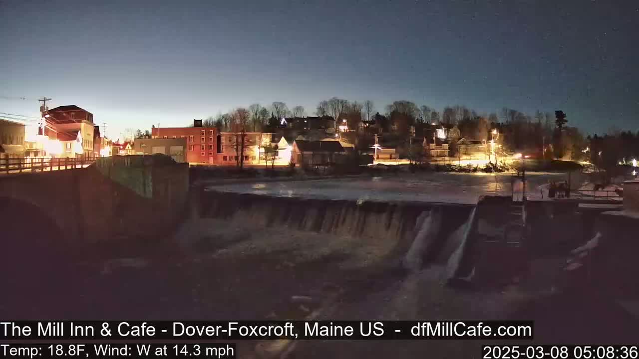 A nighttime view of a town's illuminated buildings and a partially frozen river flowing over a dam, under a mostly clear, dark sky.