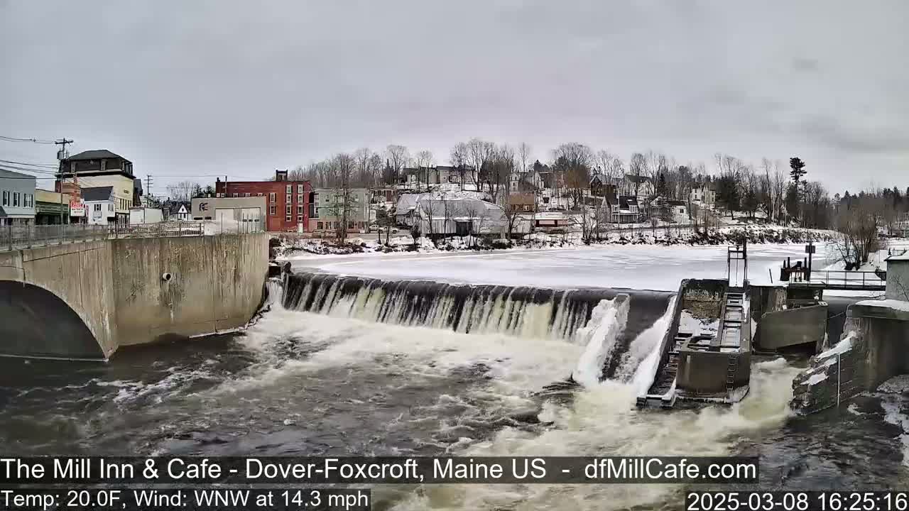 A partially frozen river flows over a dam in a town, under an overcast sky.