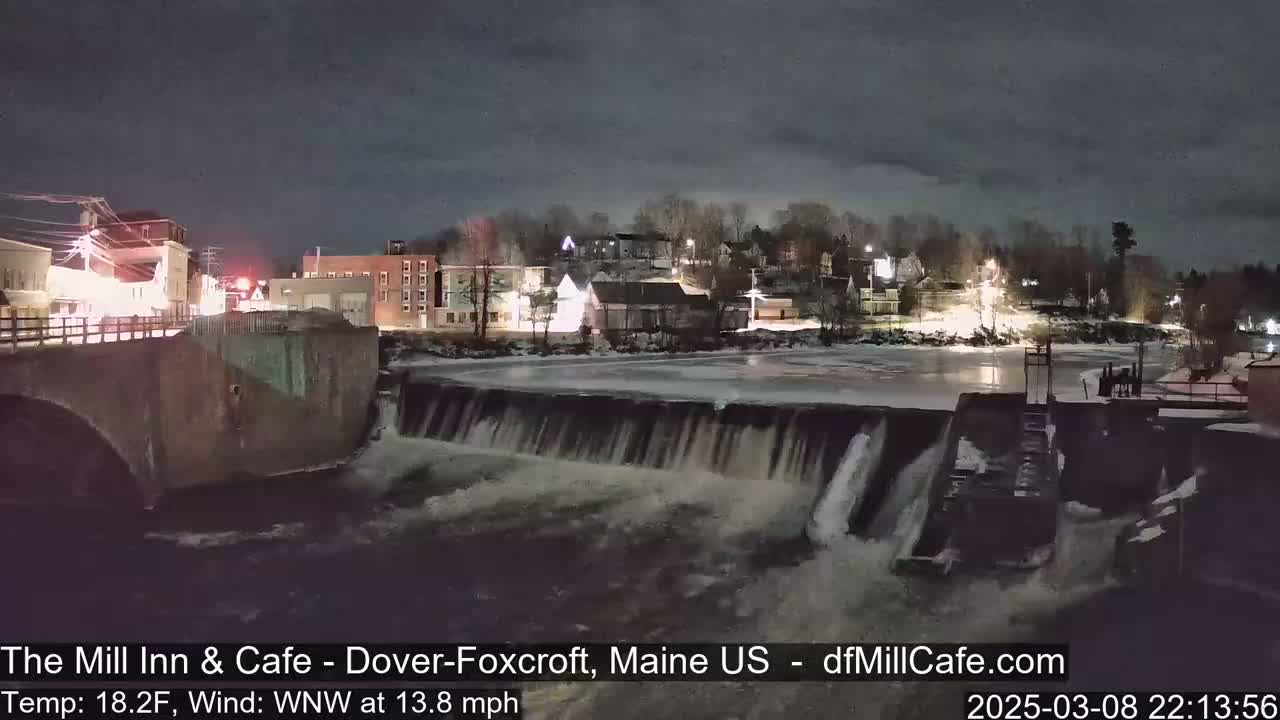 A nighttime view of a town's illuminated buildings and a dam with water flowing over it, under a dark, cloudy sky.
