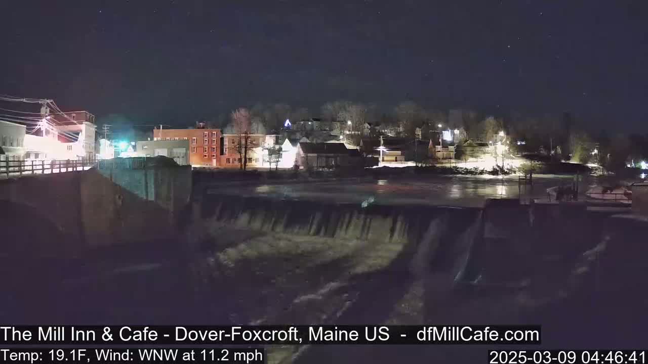A nighttime view of a town's waterfront, featuring a dam with flowing water under a clear, starry sky.