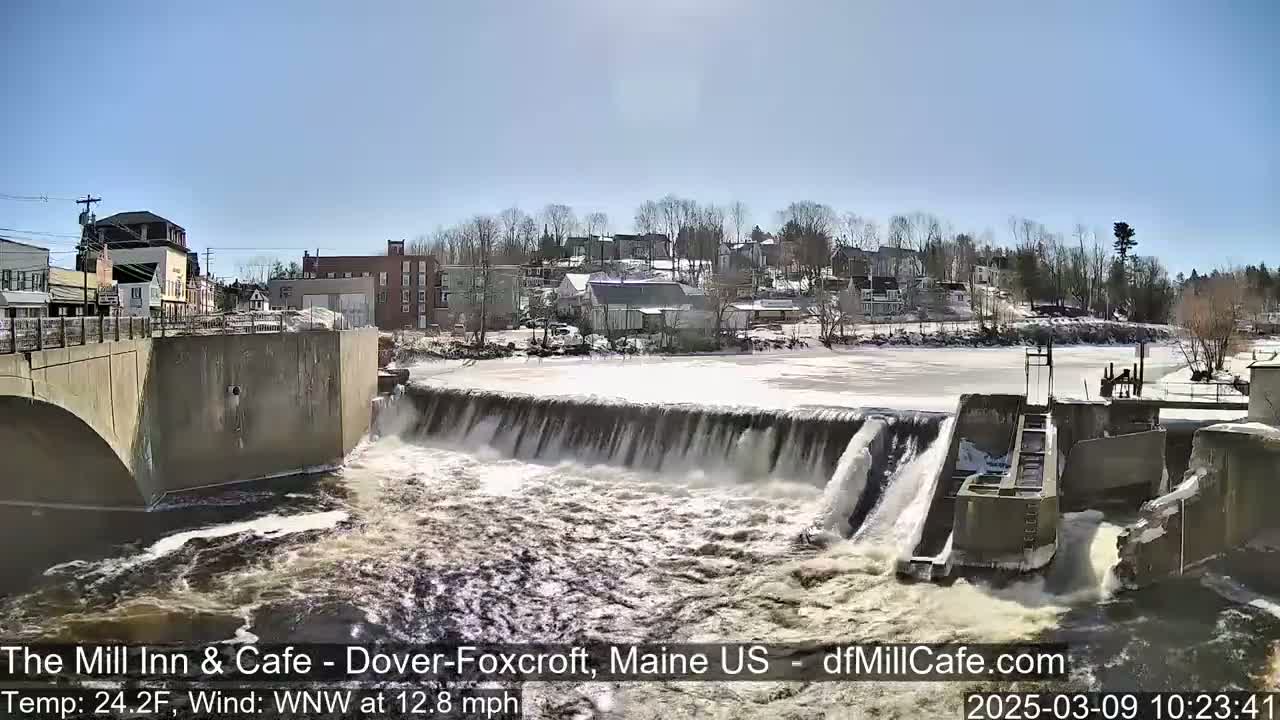 A dam with water flowing over it in a town on a sunny day with a temperature of 24.2°F and a wind of 12.8 mph from the west-northwest.