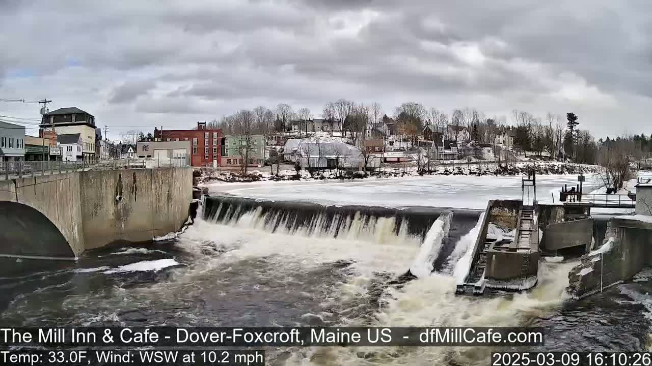 A river flows over a dam in a town under an overcast sky.