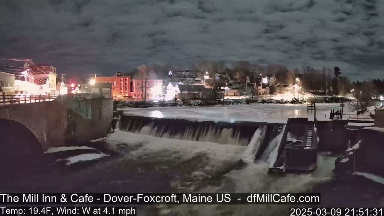 A nighttime view of a dam and river flowing through a small town, with partially frozen water and a cloudy sky.