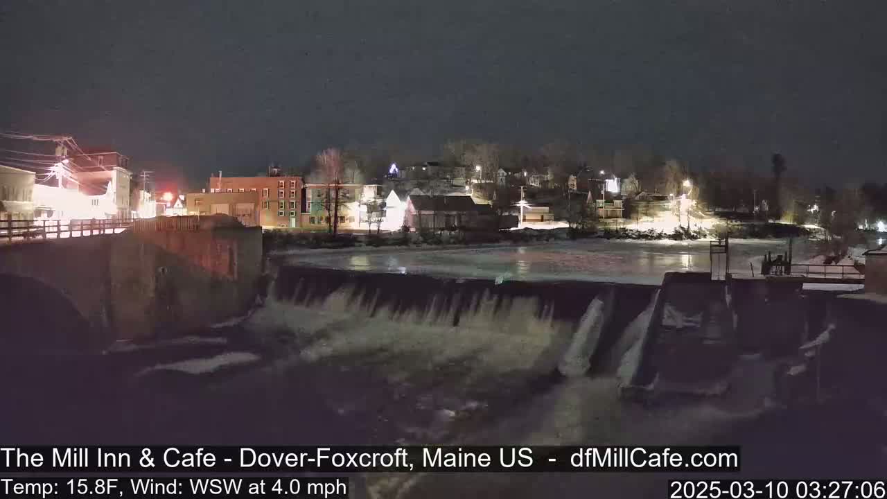 A nighttime view of a town's illuminated buildings and a dam with flowing water over it, partially frozen river, under a dark sky.