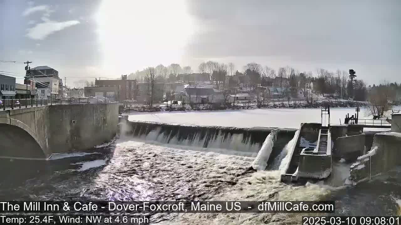 A dam with flowing water partially frozen over, situated in a small town under a partly sunny sky.