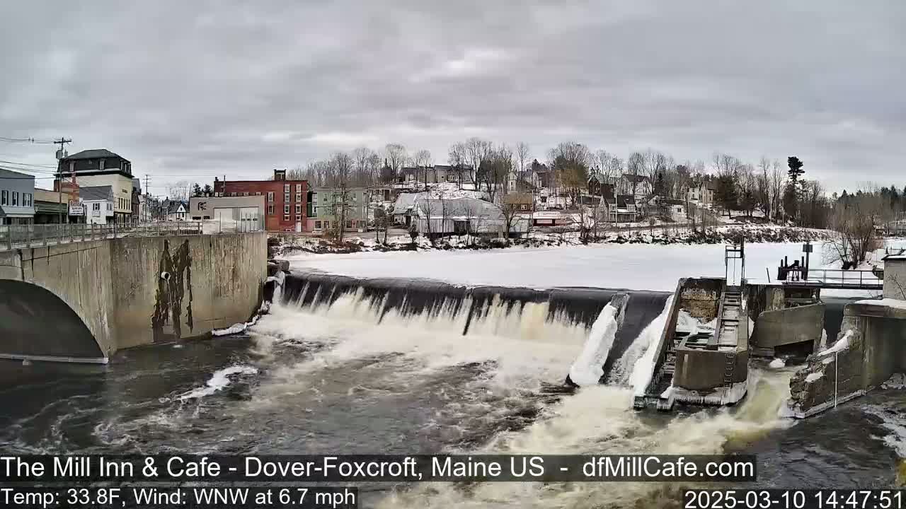 A river flows over a dam in a town under an overcast sky, with snow and ice visible on the river and surrounding areas.