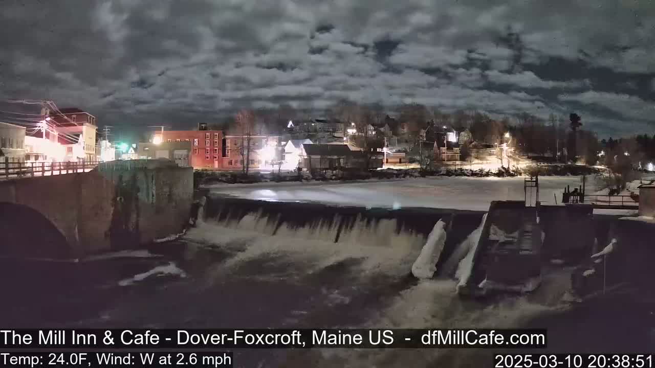 A nighttime view of a town's illuminated buildings and a dam with flowing water under a cloudy sky.