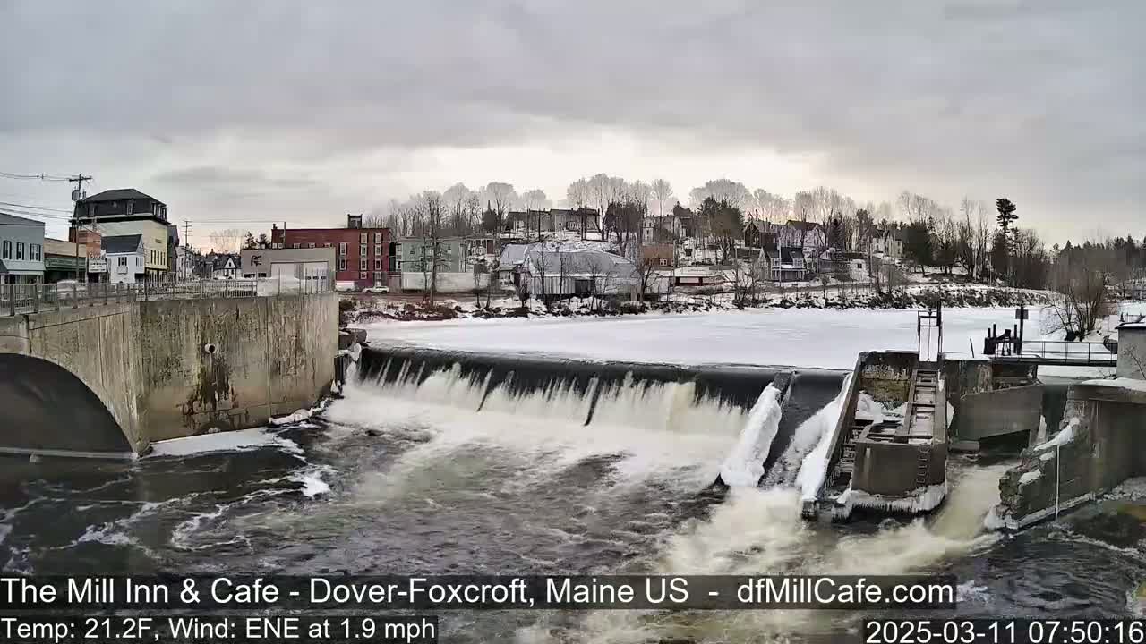 A partially frozen river flows over a dam in a town with buildings lining the riverbank under an overcast sky.
