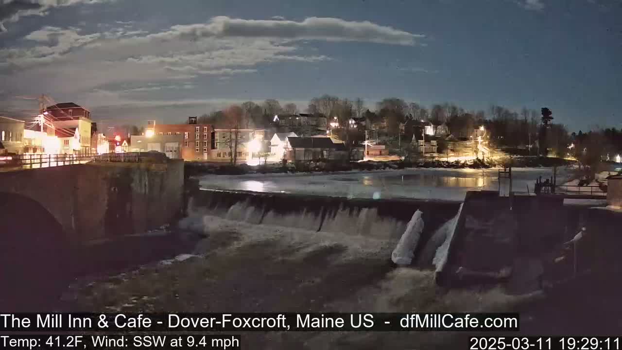 A nighttime view of a town's illuminated buildings and a dam with flowing water under a partly cloudy sky, with a temperature of 41.2 degrees Fahrenheit and a south-southwest wind at 9.4 mph.