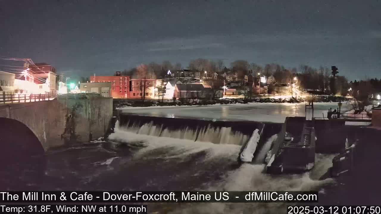A nighttime view of a dam with water flowing over it, partially frozen river, and a small town lit up in the background under a clear, starry sky.
