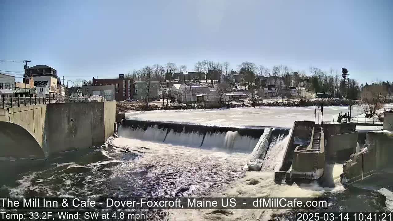 A river flows over a dam in a town on a partly sunny day with a temperature of 33.2°F and a southwest wind.