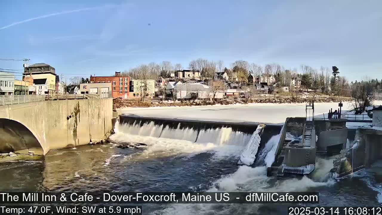 A river flows over a dam in a town on a sunny day, with some ice remaining on the river upstream from the dam.