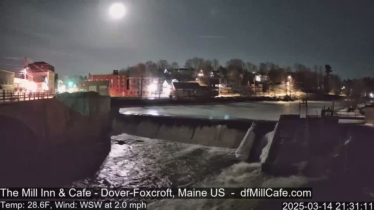 A brightly lit town at night is seen from above a dam, with a full moon in the clear sky and a river flowing over the dam.