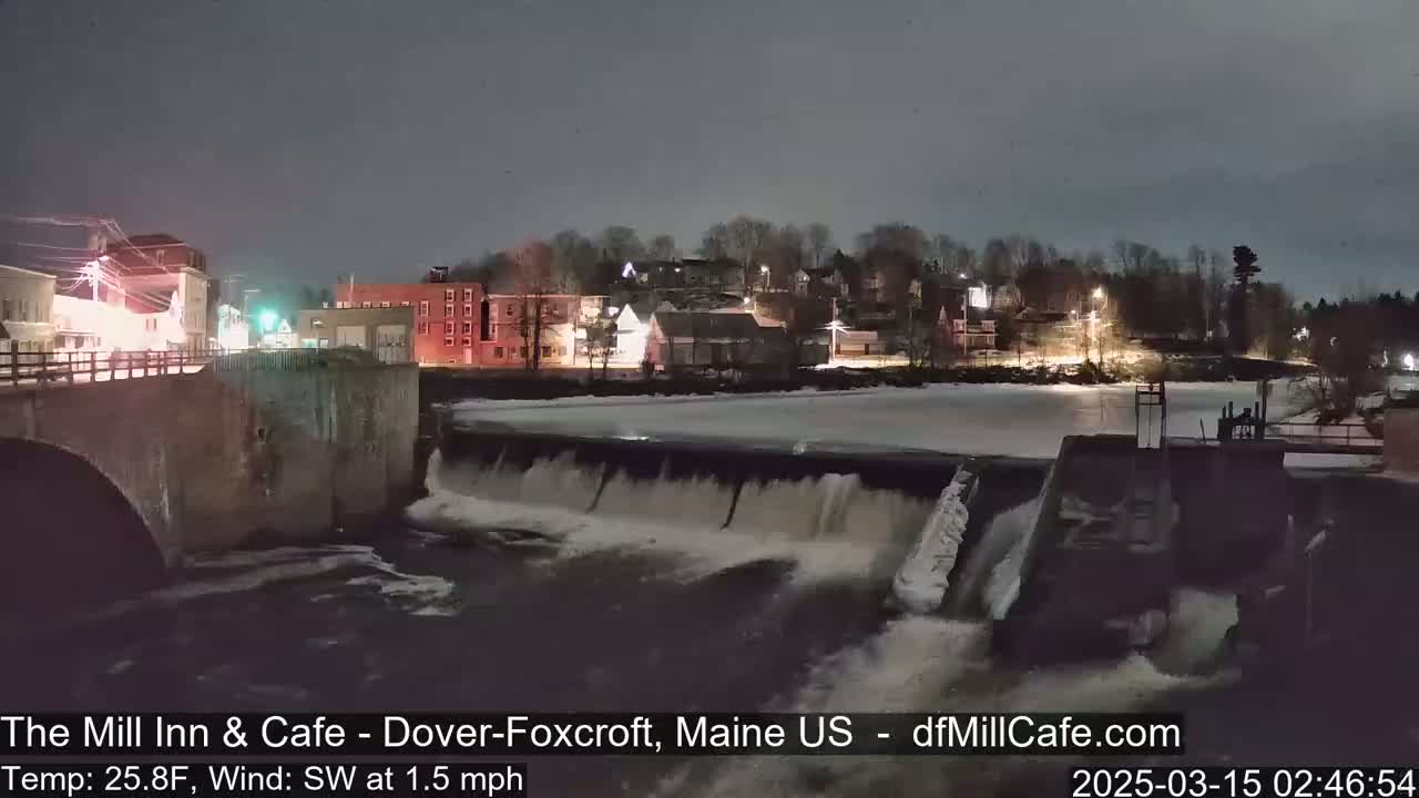 A nighttime view of a dam with water flowing over it, illuminated buildings in the background, and a partially frozen river under a dark sky.